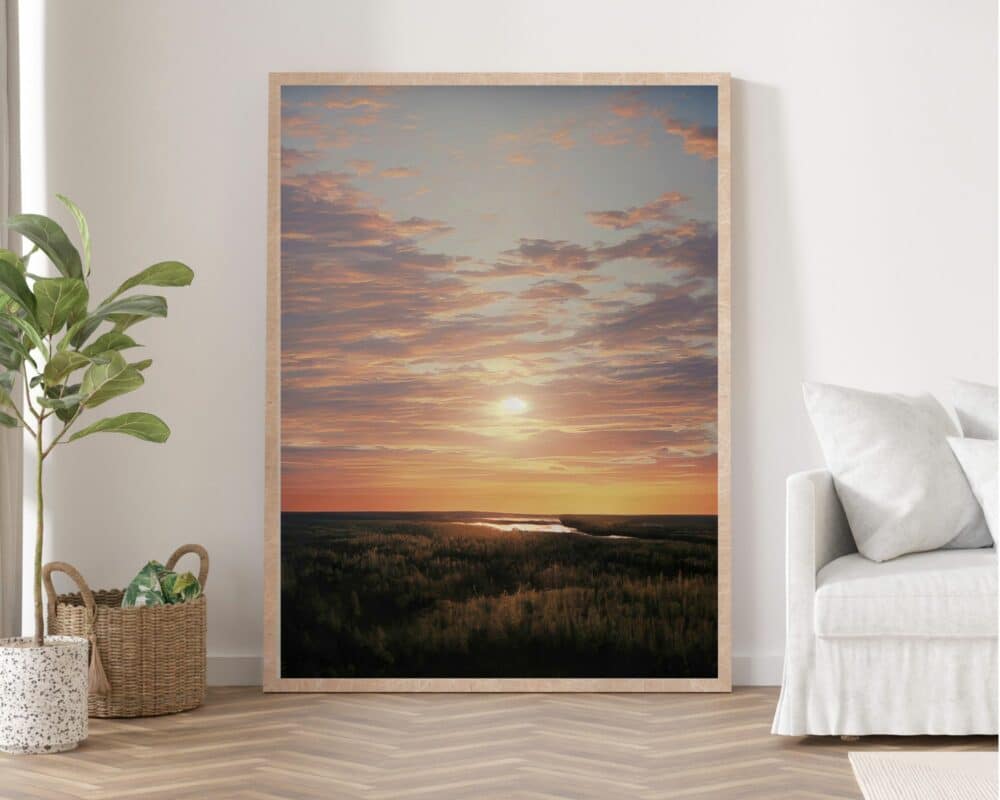 **תמונה לחדר שינה ממוסגרת נוסח ציור וינטג' שקיעה מעל יער** מונחת על קיר לבן. עציץ ירוק יושב משמאלו, וסלסלה ארוגה מלאה במגזינים נמצאת על הרצפה לידו. מימין יש ספה לבנה עם שמיכת זרקה אפורה בהירה.