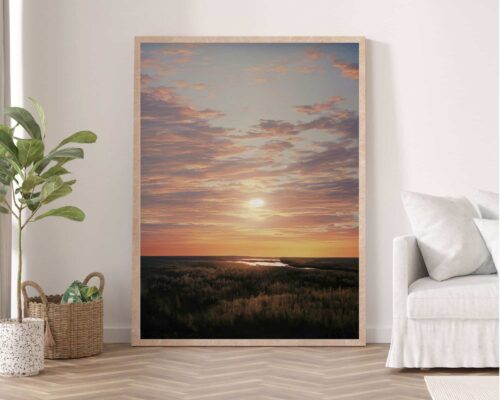 **תמונה לחדר שינה ממוסגרת נוסח ציור וינטג' שקיעה מעל יער** מונחת על קיר לבן. עציץ ירוק יושב משמאלו, וסלסלה ארוגה מלאה במגזינים נמצאת על הרצפה לידו. מימין יש ספה לבנה עם שמיכת זרקה אפורה בהירה.