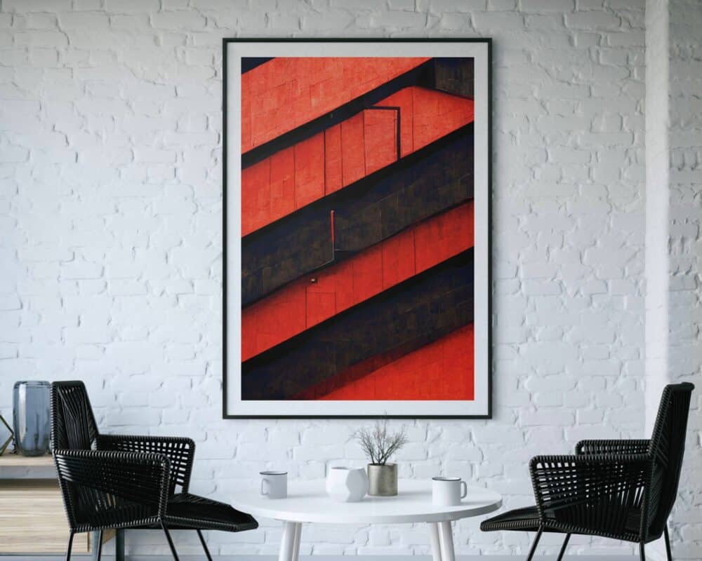 יצירת אמנות בעלת מסגרת גדולה, המכונה תמונה למשרד בעיצוב אבסטרקט קווים אדום ושחור, המתארת חזית בניין מודרנית ומופשטת בצבע אדום ושחור, תלויה על קיר לבנים לבנים. האמנות מרוכזת מעל שולחן לבן עגול קטן עם שני כסאות נצרים שחורים משני הצדדים. העיצוב המינימליסטי כולל צמח קטן במרכז השולחן.