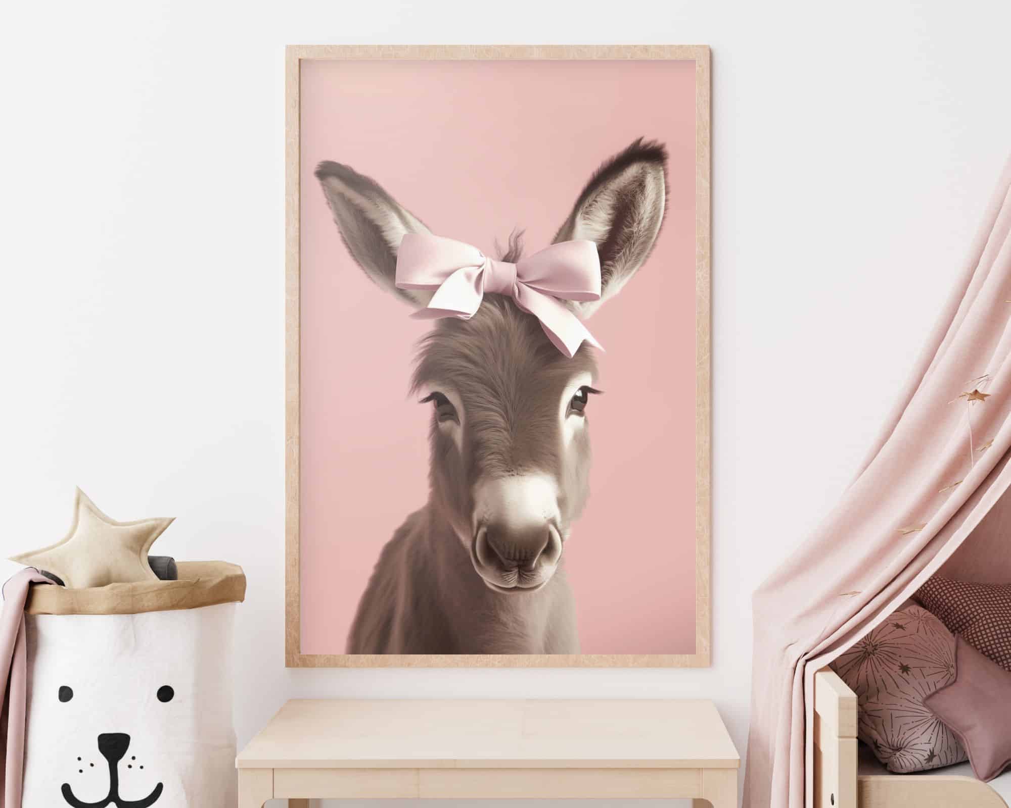9272mo1 תמונה ממוסגרת שכותרתה "חמור בייבי (עיר) עם פפיון בורוד עדין" תלויה על קיר לבן. מתחת לתמונה שולחן עץ בהיר. משמאל לשולחן יש סל אחסון לבן בעיצוב פנים דובי, מלא בצעצועי כוכב קטיפה וירח.