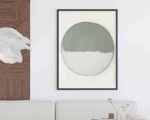 ציור מינימליסטי שכותרתו "תמונה לחדר שינה נוסח מינימליסטי עיגול לבן וירוק מרווה" במסגרת שחורה תלוי על קיר לבן. הגרפיקה כוללת עיגול גדול וממורכז כאשר החצי העליון בצבע אפור והחצי התחתון בלבן. מתחת לציור ספה לבנה עם שולחן צד מעץ ובו פרחים מיובשים. משמאל, יש קטע של חיפויי עץ עם מרקם ויצירת נייר לבן ומקומט.