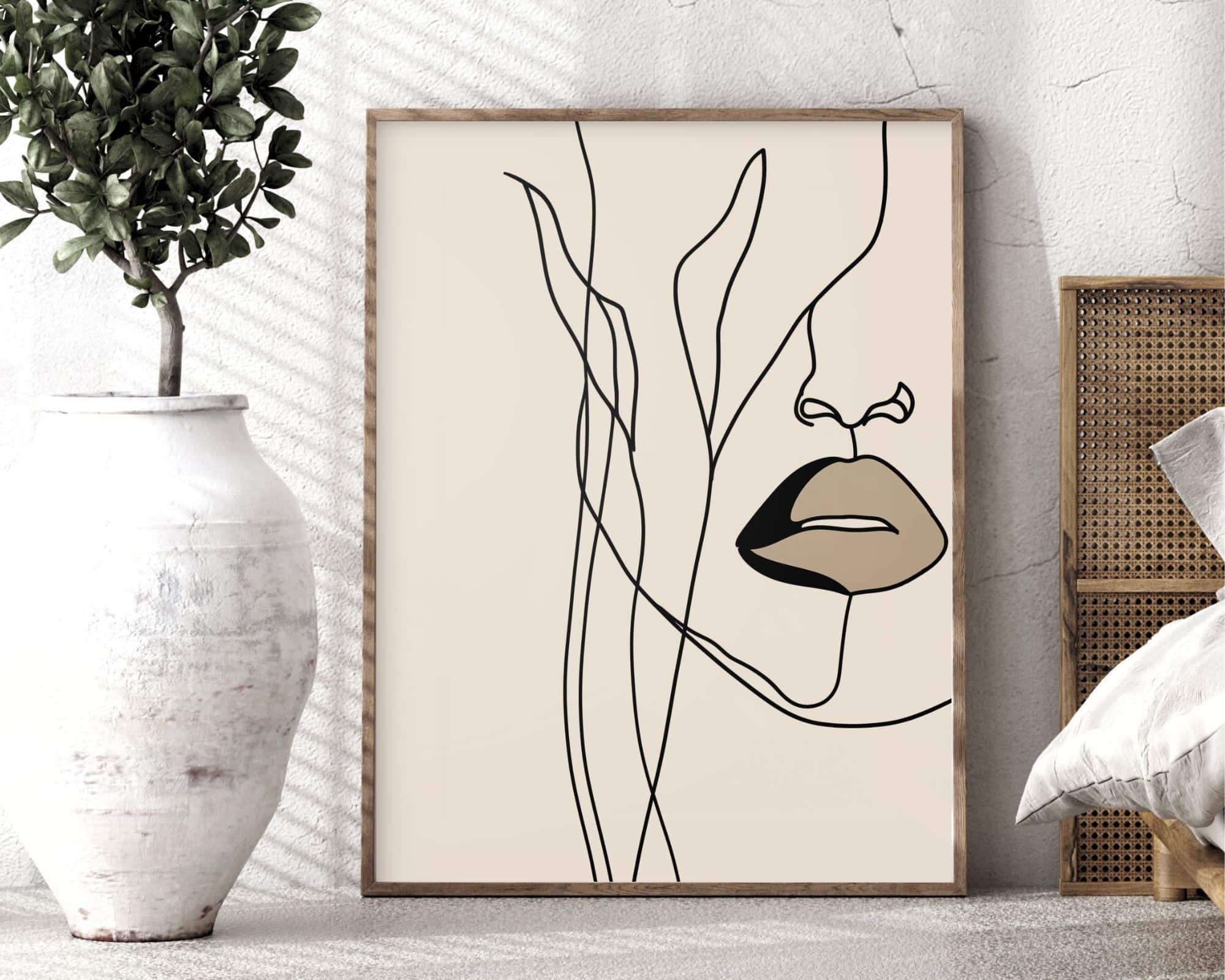 2574mo1 רישום מינימליסטי הכולל פנים מופשטות וחלקיות עם דגש על השפתיים והאף מוצג במסגרת עץ. יצירת האמנות, בשם "תמונה לחדר שינה בעיצוב מינימליסטי ציור קווי אישה ועלים צבע בז'," מונחת על הרצפה לצד שולחן צד נצרים וליד אגרטל לבן גדול עם עלים ירוקים.