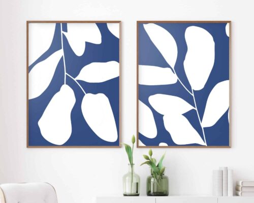 שני הדפסים ממוסגרים מקולקציית **סט פוסטרים לסלון מעל הספה נוסח מינימליסטי עלים בכחול לבן**, הכוללים צלליות עלים לבנים על רקע כחול, מותקנות על קיר לבן. מתחת להדפסים, ארון לבן מחזיק שני אגרטלי זכוכית עם סידורי פרחים מינימליסטיים, המדגישים את העיצוב הנקי והמודרני.