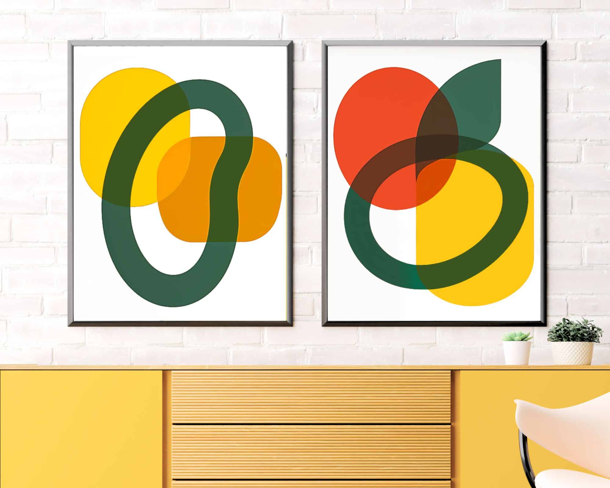 2606mo1 שני הדפסים ממוסגרים מסט סט פוסטרים למטבח נוסח מינימליסטי פירות מופשט תלויים על קיר לבנים לבנים מעל שידה צהובה. ההדפס השמאלי כולל צורות חופפות של ירוק, צהוב וכתום. ההדפס הימני מציג צורות מצטלבות באדום, ירוק, צהוב וכתום. קקטוס קטן יושב על השידה.