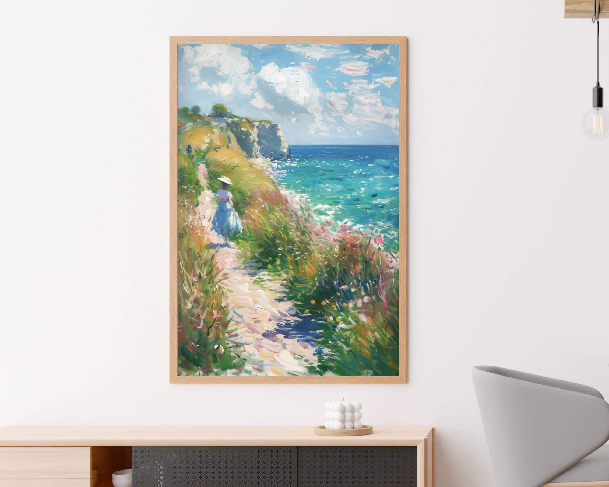 2615mo1 תמונה ממוסגרת לפינת אוכל נוסח ציור וינטג' אישה בשדה פורח תלויה על קיר לבן מעל מזנון עץ. התמונה מתארת אישה מטיילת לאורך שביל עטור פרחים ליד האוקיינוס, עם צוקים רחוקים ושמים בהירים ומעוננים. בקרבת מקום, יש כיסא מודרני ונורה תלויה.