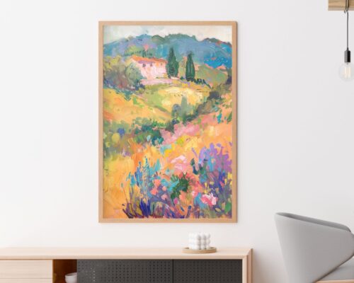 תמונה ממוסגרת שכותרתה "תמונה לפינת אוכל ציור וינטג' בשדה צבעוני" תלויה על קיר לבן. הסצנה מציגה בית השוכן בין גבעות עם עצים, שיחים צבעוניים ופרחים בחזית. גוף תאורה מודרני מינימליסטי וכיסא אפור נראים חלקית בצדדים.