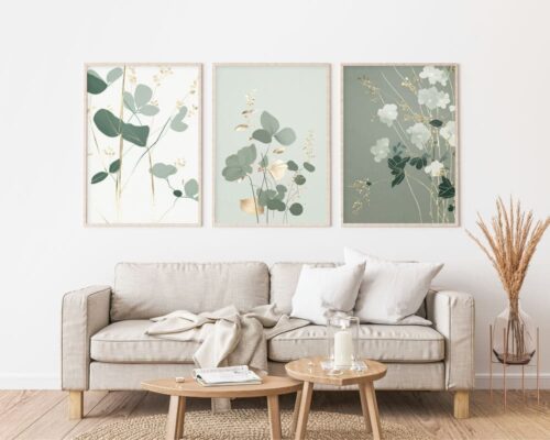 סלון נעים עם ספה בז' מעוטרת בכריות לבנות וזריקה קלה. מעל הספה מונחים על הקיר שלושה הדפסי אמנות בוטניים ממוסגרים מתוך סט "סט 3 תמונות לסלון נוסח מינימליסטי צמחים ירוק עדין". שולחן קפה מעץ מלפנים מכיל נר, ספר ואגרטל. עשבים מיובשים באגרטל עומד ליד הספה.