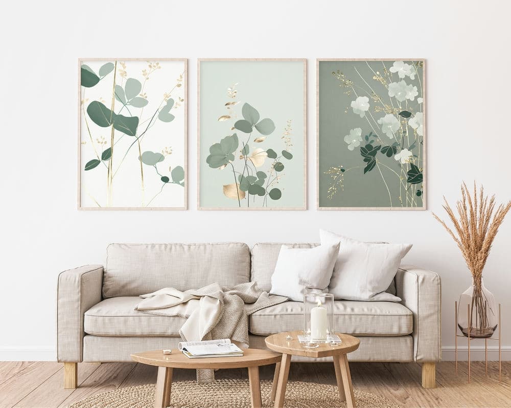 סלון נעים עם ספה בז' מעוטרת בכריות לבנות וזריקה קלה. מעל הספה מונחים על הקיר שלושה הדפסי אמנות בוטניים ממוסגרים מתוך סט "סט 3 תמונות לסלון נוסח מינימליסטי צמחים ירוק עדין". שולחן קפה מעץ מלפנים מכיל נר, ספר ואגרטל. עשבים מיובשים באגרטל עומד ליד הספה.