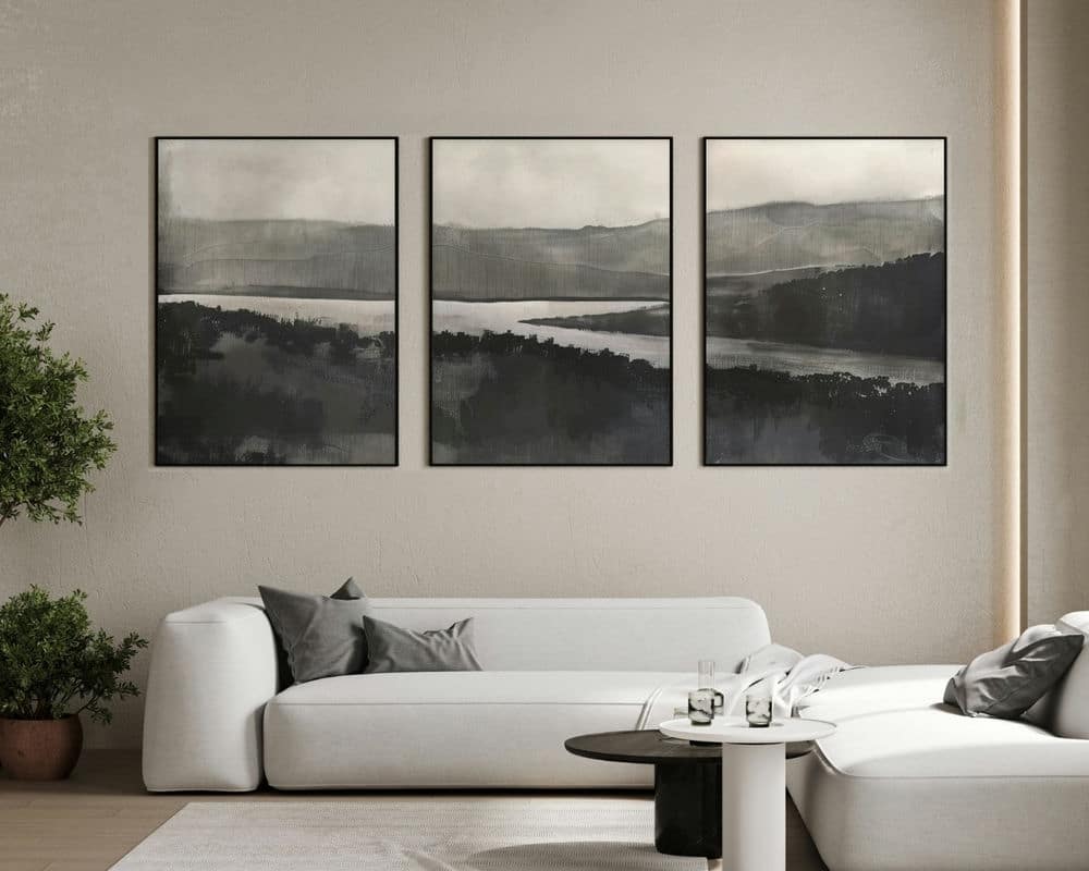 סלון מודרני כולל ספת חתך לבנה עם כריות אפורות, שולחן קפה עגול עם כלי זכוכית ועציץ. מעל הספה תלויים שלושה ציורי נוף מופשטים ממוסגרים של נהר ביער בסגנון שחור ולבן מינימליסטי מתוך הסט סט 3 תמונות של נהר ביער שחור לבן לסלון נוסח מינימליסטי תלויים על הקיר בז'.