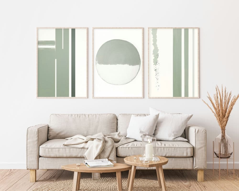 סלון מינימליסטי כולל ספה בצבע בז' בהיר המעוטרת בכריות לבנות ובז'. מעל תלויה על הקיר סט 3 תמונות לסלון נוסח מינימליסטי קווים ירוק עדין, המציגה שלוש יצירות אמנות מופשטות בגווני ירוק ולבן עמומים. החדר כולל גם שולחן קפה מעץ עם נרות ואגרטל עם צמחים יבשים.