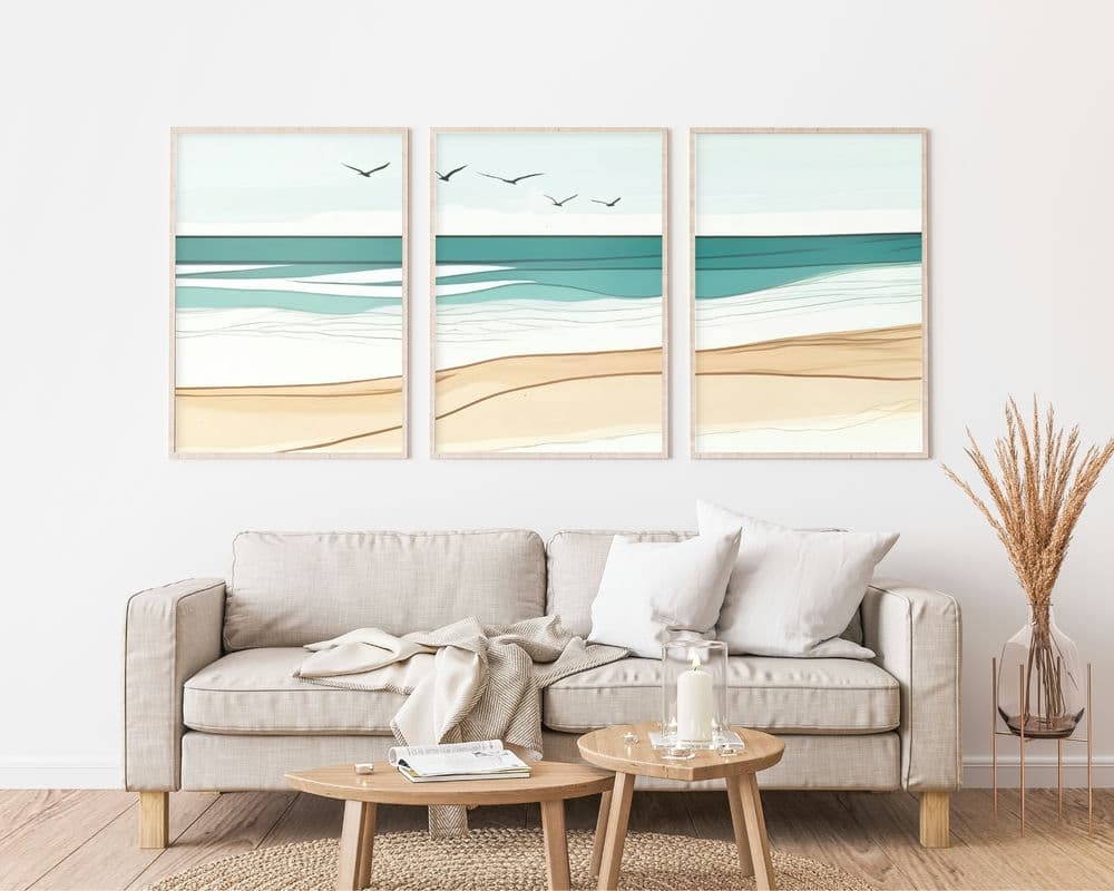 סלון מינימליסטי כולל ספה בצבע בז' עם כריות לבנות ואפורות. שולחן קפה מעץ מכיל ספרים, נרות וכוס. מעל הספה תלוי על הקיר טריפטיך של אמנות חוף בוהמית מקולקציית סט 3 תמונות לסלון נוסח בוהו חוף ים רגוע, המציג גווני טורקיז, בז' ולבן. אגרטל עם חיטה מיובשת מקשט את הצד הימני.