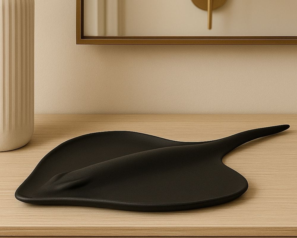 A מעמד שולחני לתכשיטים בצורת מנטה ריי, בעיצוב שחור ואלגנטי, מוצב על משטח עץ בהיר לצד אגרטל קרמי חרוץ בגוון קרם, עם מראה וקיר ניטרלי ברקע.