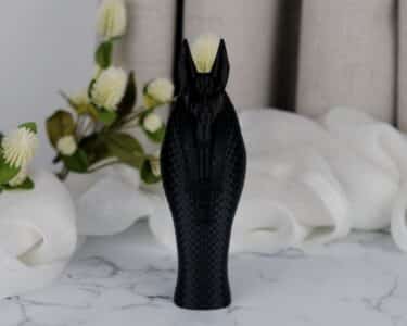 פסל אנוביס – אל המוות המצרי, שחור ובעל מרקם, ניצב על משטח שיש, עם בד לבן ופרחים בגווני קרם ברקע.