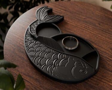 מגש תכשיטים בצורת דג קוי שחור עם קשקשים מעוצבים מונח על שולחן עץ, עליו טבעת עדינה עם אבנים קטנות. מסביב נראות עלים ירוקים.