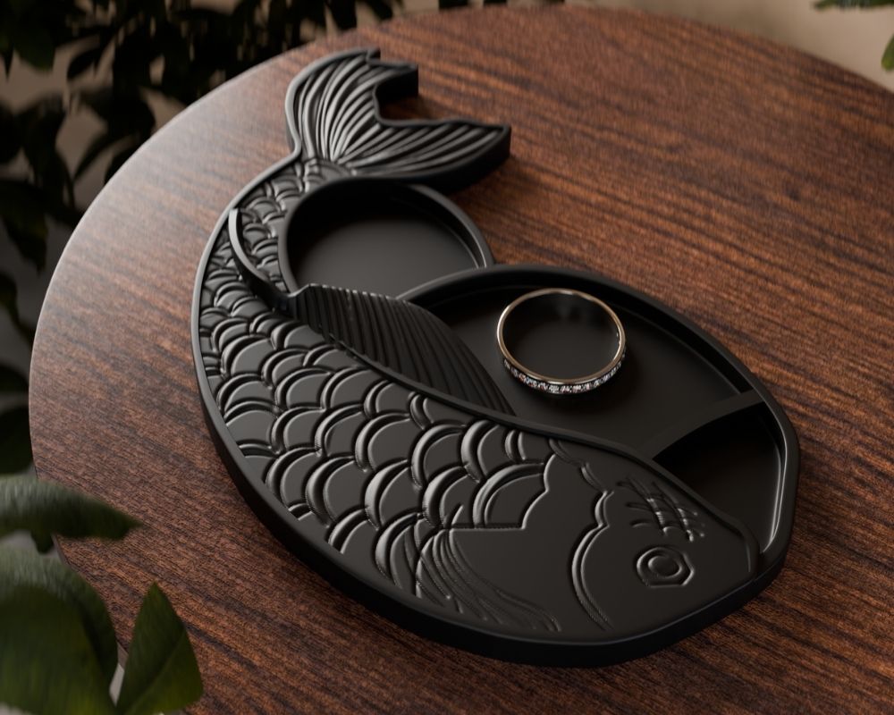 מגש תכשיטים בצורת דג קוי שחור עם קשקשים מעוצבים מונח על שולחן עץ, עליו טבעת עדינה עם אבנים קטנות. מסביב נראות עלים ירוקים.