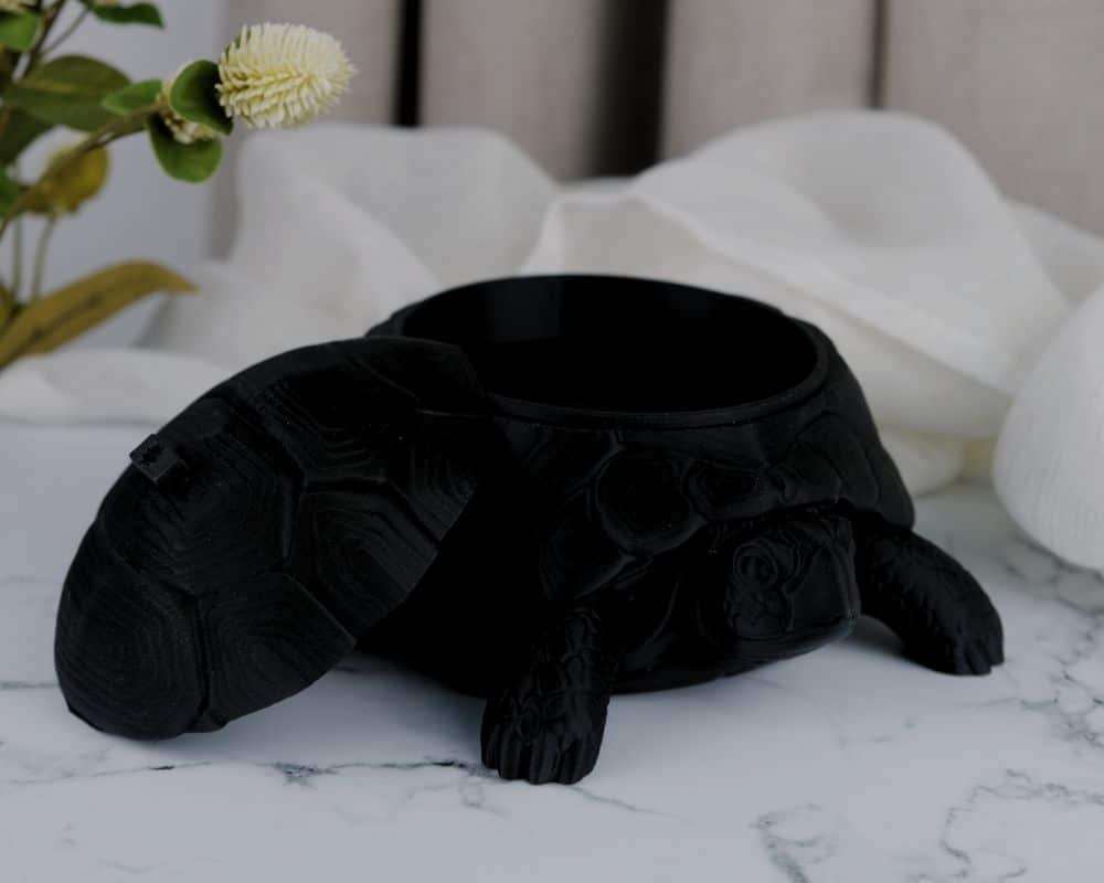 קופסת תכשיטים בצורת צב עם מכסה קונכיה פתוח מונחת על משטח שיש, ליד בד לבן וצמח ירוק ברקע.