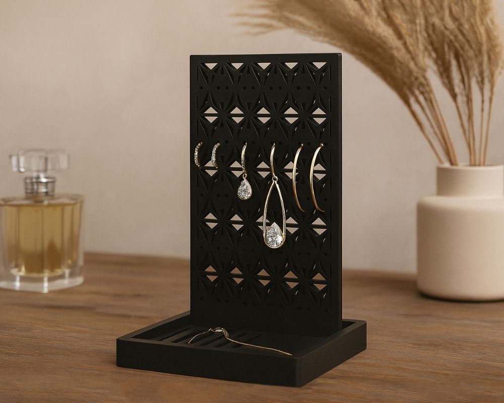 מעמד עגילים גאומטרי שחור מציג זוגות עגילים כמו חישוקים ודנגלים על משטח עץ. ברקע המטושטש נראים בקבוק בושם ואגרטל עם עשב יבש.