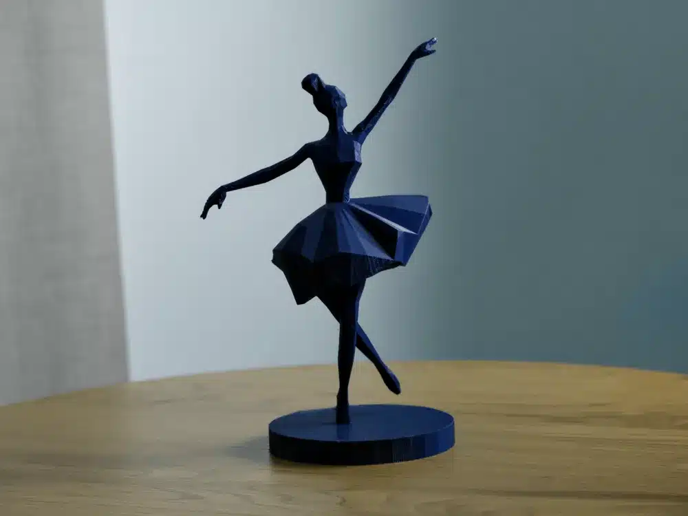 2025-01-15_7ca0ba03a9199 פסלון בלרינה גיאומטרי בצבע כחול כהה עומד על רגל אחת על גבי בסיס עגול, בתנוחה של זרוע אחת מורמת והשנייה מושטת, מוצג על משטח עץ על רקע מואר בעדינות.