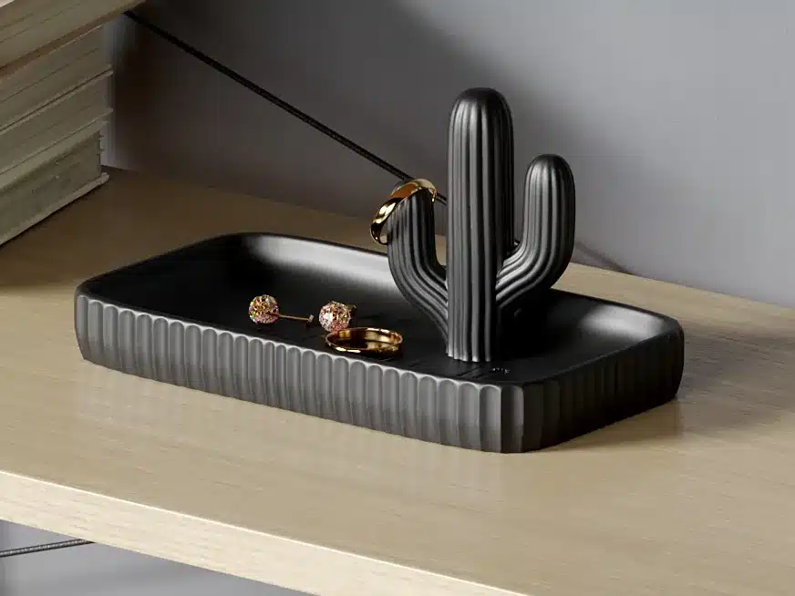 מגש שחור ומלבני עם מחזיק טבעות בצורת קקטוס מונח על מדף עץ בהיר, ועליו מונחים טבעת זהב, זוג עגילים עם אבני חן צבעוניות וטבעת זהב נוספת התלויה על הקקטוס.