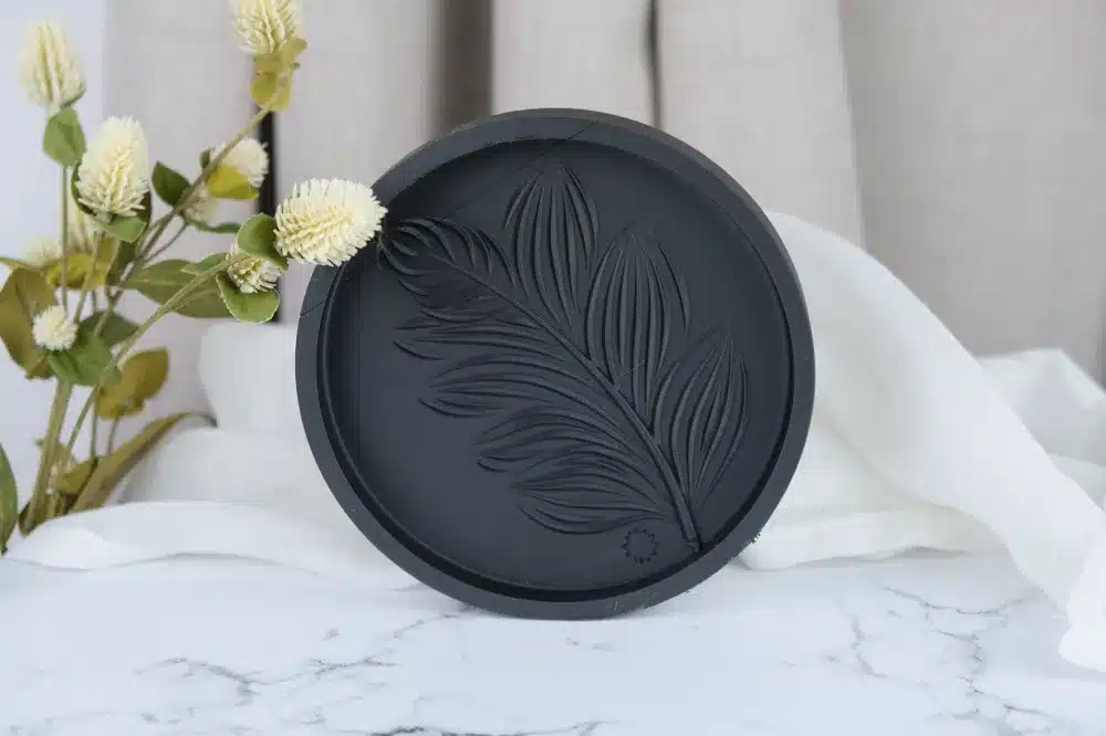 סבון שחור עגול עם עיצוב עלים בולט מונח על משטח שיש לבן, לצד ענף פרחים צהובים ולבנים ורקע מבד לבן.