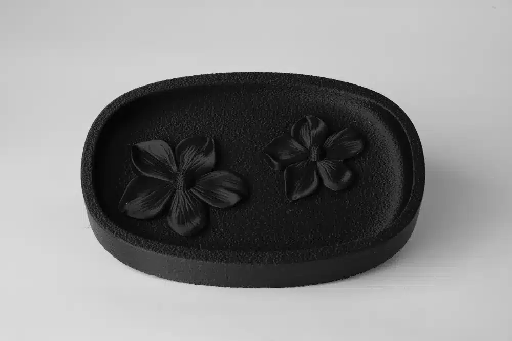 סבונייה שחורה אלגנטית בצורת אליפסה בשם מגש פרחים אלגנטי, עם משטח מרקם ושני עיצובים של פרחים שחורים, המוצגת על רקע בהיר וחלק.