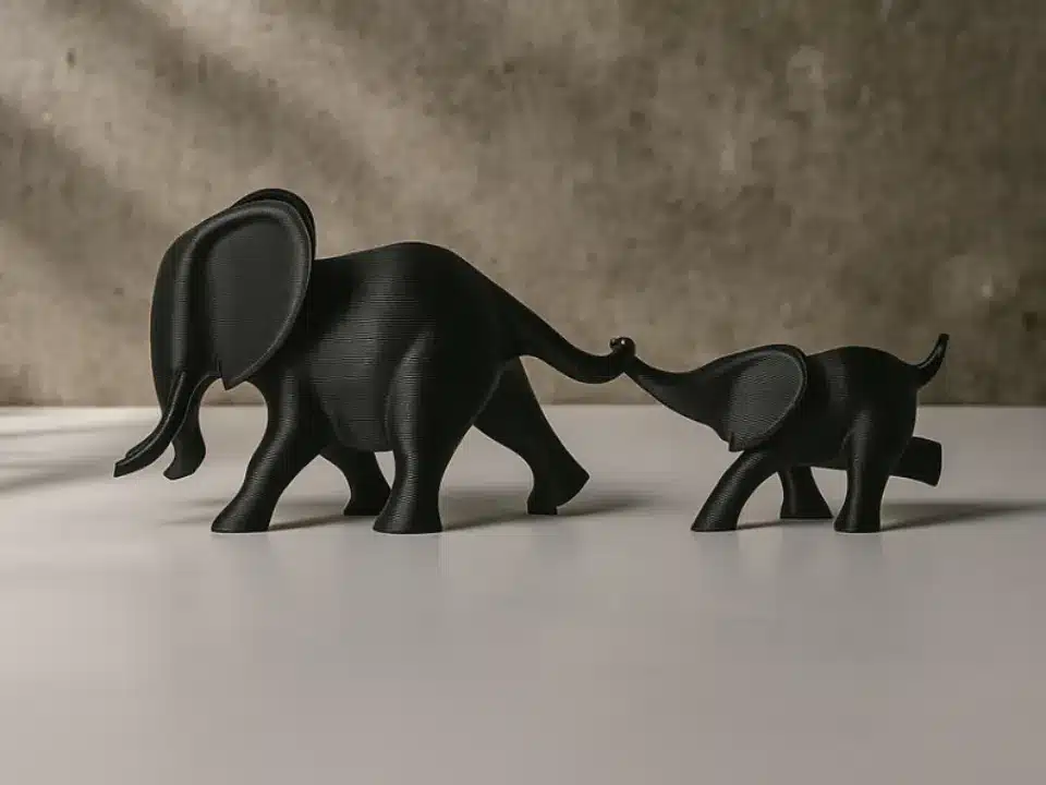 2025-09-17_6027919e880a5 שתי פסלונים מינימליסטיים של פילים שחורים, אחד גדול ואחד קטן, עומדים על משטח לבן. הפיל הקטן אוחז בזנבו של הפיל הגדול יותר באמצעות חדקו. הרקע הוא קיר ניטרלי ומחוספס.